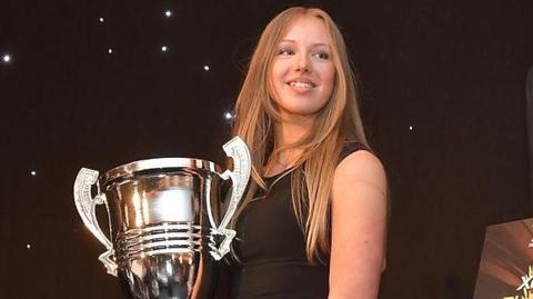 A girl with long hair wearing an evening dress holding a large trophy
