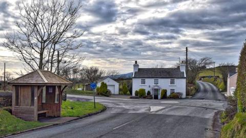 The Braaid crossroads, with a bus stop on the left and a white cottage in the centre.
