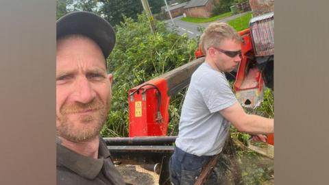 A selfie taken by Daniel Graham shows Daniel Graham in the foreground and Adam Carruthers working behind him. Daniel Graham is looking at the camera and wearing a black cap and black shirt. Adam Carruthers is side on and wearing sunglasses and a grey top