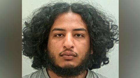 Qusai Abdalla's mugshot. He has a dark beard, brown eyes and shoulder-length, bushy dark hair with a central parting. He is wearing a grey t-shirt and is in front of a plain grey background