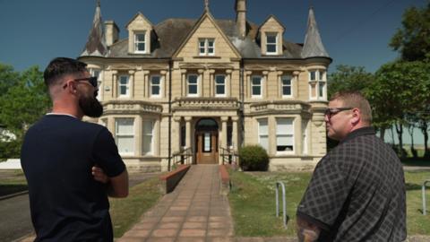 Dan Stanaway and David Bishop stand in front of the grand Victorian school building. The three-storey, stone, double-fronted building has a portico entrance with bay windows either side and small pointed turrets on the corners of the roof.