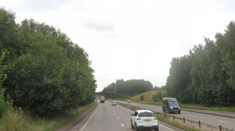 The A50 eastbound in Derbyshire, with a white car at the front of the image and trees either side of the road