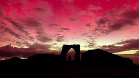Grosnez Castle in dark lighting with a pink and yellow sky above it and a person stood in the archway.