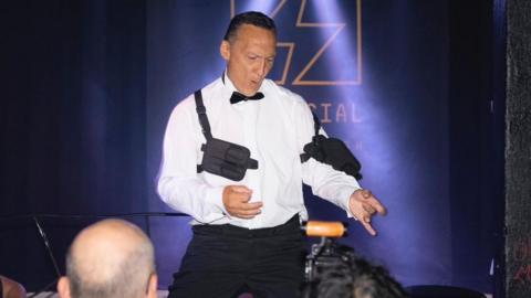 Piers Burnell is wearing evening dress, minus the blazer, whilst air guitaring in front of a crowd.