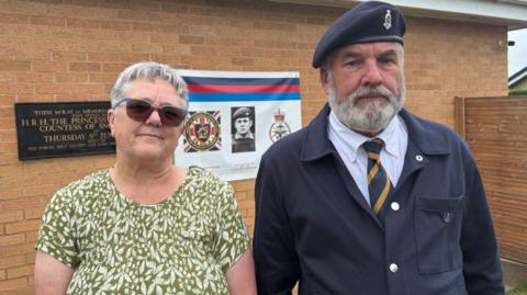 An elderly man and woman stand together infront of military plaques on a wall. She is dressed in a green and white flowery dress and he is navy blue military uniform with beret.