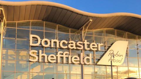 A sign on the front of the airport building reads Doncaster Sheffield Robin Hood. The white sign is on a glass wall. The building has a grey wavy roof.