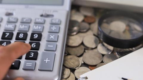 Finger moving towards a calculator next to coins, a notepad, pencil and a magnifying glass