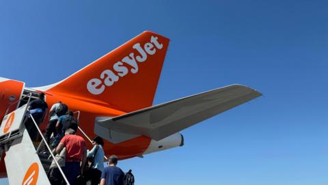 People queuing on the stairs to enter from the back of an Easyjet plane. Blue clear skies behind.