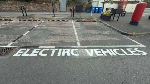 Two electric vehicle bays with misspelt road markings reading "electirc vehicles" at the side of a residential street. A traditional British phonebox and letterbox can be seen on the pavement.