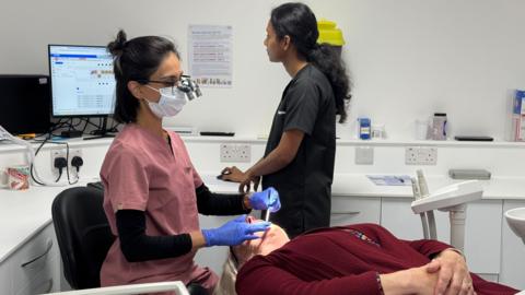 A woman in a dentist chair being examined by a dentist to her left. Behind them, a dental assistant wearing grey looks at a computer screen