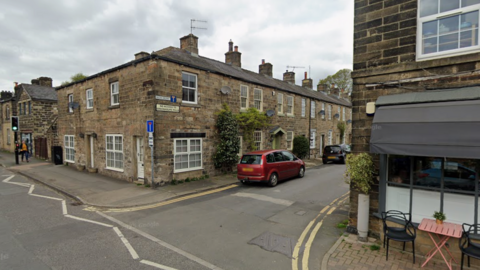 A quiet village street. On the right is a cafe awning with chairs outside. On the left is a pedestrian crossing