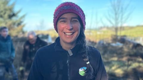 Anne smiling at the camera as she stands in a wooded area. She is wearing a black zip-up fleece with an RSPB badge, a pink and blue hat, and has her dark brown hair in a plait.