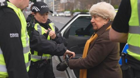 A woman, wearing a brown jacket and cream-coloured puffy hat, is arrested by a female officer next to a police car. Two other officer stand on either side of them in the foreground.
