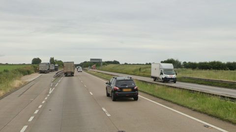 The westbound carriageway of the A180. The beige concrete surface can be seen alongside a green, grass verge and a metal barrier. A black car is driving along the road near a lorry.