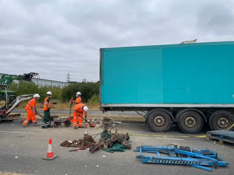 Workers in hi-vis clothing clearing the site. There are metal items laid out on the floor to the side of a blue trailer.