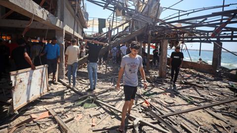 A view of the destruction after air strikes on a coffee shop in Gaza. People are pictured examining the debris with a young man walking out of the cafe in the foreground, and the sea and blue sky in the background, taken on 30 June.