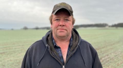 Dan Willis stands in a field at his farm. He is wearing a khaki coloured cap and a check blue and navy shirt under a zipped hooded jumper. The field behind him is tinted white with frost.
