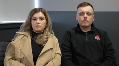 A man and woman sit together looking at the camera, the woman is wearing a camel coloured coat and her partner a black sweatshirt.