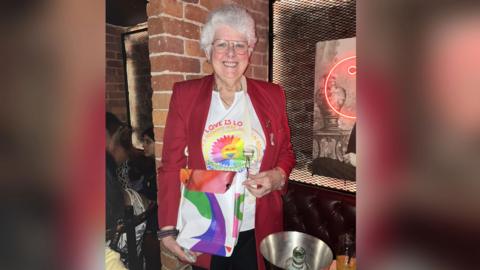 Judith Whiston, a woman wearing glasses, a white T-shirt and red jacket, is standing in a bar. She is smiling as she poses for the photo.