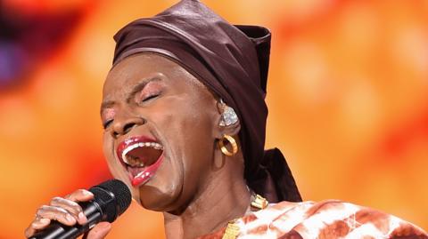 Angélique Kidjo sings into a microphone with her eyes closed. She wears gold hoop earrings, red lipstick and a brown headscarf.