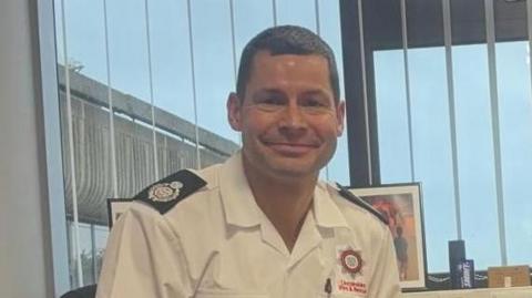 Joe Haustead smiling at the camera. He is wearing uniform. Behind him is a window with blinds and a view of blue skies.