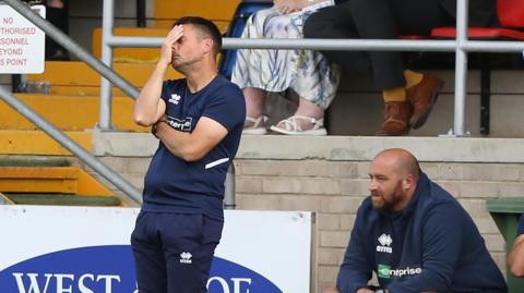 A wide shot of Jimmy Ball with his face in his hands after his side have made a mistake. He has short dark hair and is wearing a navy blue training t-shirt and tracksuit bottoms.