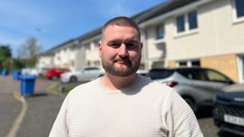 A man in a white tshirt with short brown hair and a beard stands in a residential street