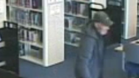A blurred CCTV image of a man who is walking through the library. He is wearing a black hooded coat and a dark grey or green cap. There are a number of library shelves behind him, as well as a library table. 