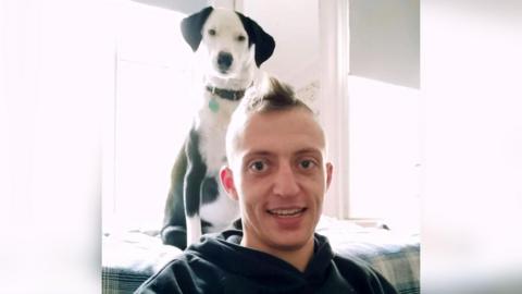 Matty looks at the camera with a small black and white dog sitting on the back of the sofa behind them. They are smiling at the camera and have a black hoodie on