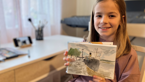 Image of Kevin who is sitting on a wooden chair and wearing a pick t-shirt. He has shoulder length blonde hair. He is holding his watercolour art up to the camera and smiling. The art is of a steam ship in the water, docked up.