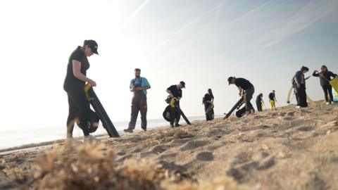 Multiple people on a beach using vacuums, viewed from ground level with sky in background.