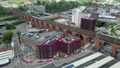 A drone image of Stockport town centre. There is a train travelling along the historic viaduct which spans the length of the picture. In the middle of a ring road is a number of new, modern high rises located next to former mill buildings.