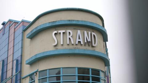 A teal and light yellow building has the white letter 'Strand' on it