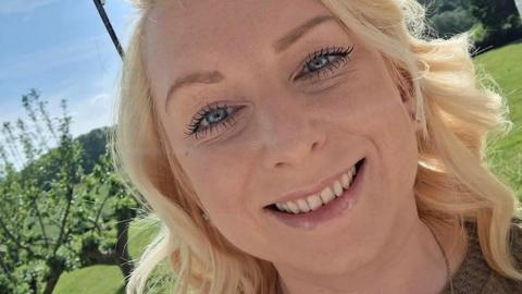 A blonde haired girl smiles at the camera. It is sunny and green in the background