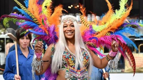 A woman wearing a carnival outfit looks at the camera with a smile. She has long platinum blonde hair and wears a multi-coloured, sequinned top with feathers of yellow, blue, orange and gold flowing up from her shoulders