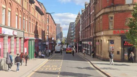 A narrow city street lined with tall red-brick buildings on both sides. There are shops and businesses at street level, with signs visible, including one that reads “DANA STORE”. Several pedestrians are walking along the pavements, and vehicles, including white vans and a red vehicle with yellow markings, are on the road. The words “BUS STOP” are painted in yellow on the street.