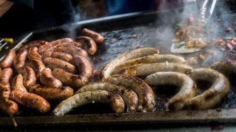 Two different varieties of sausage are being cooked on a large grill, alongside some vegetables in the background. A cook is using a spatula and some tongs. 