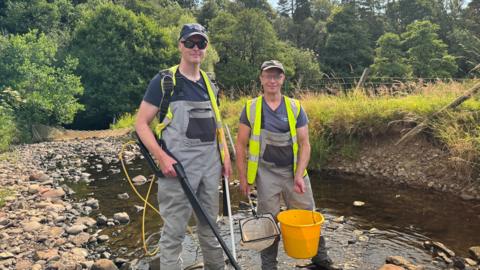Luke Bryant on the left, next to a colleague from West Cumbria Rivers Trust. They are both wearing grey overalls, blue t-shirts, hi-viz vests and baseball caps. They are holding, buckets, nets and other fishing equipment. They are standing in a shallow stream, surrounded by greenery, with trees in the background.