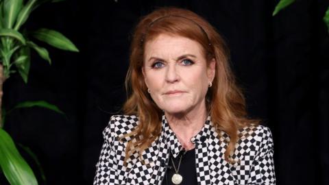 Sarah Ferguson wears a black and white checked jacket with a black top underneath, and is sitting against a black background - plants can just be seen at the side. Her red hair is pushed back with a thin black headband and she has a neutral expression on her face