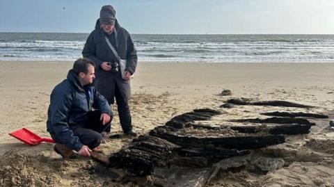 Two men investigate timbers that emerged on a beach following a storm. The remains, which just about resemble part of a ship, are blackened and covered partially by sand. One man is crouched near a red shovel. 