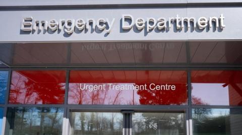 Looking up at the glass entrance to the emergency department. Above the door is a grey canopy with Emergency Department written in raised letters. Between the canopy and the top of the doors is a strip of red glass with Urgent Treatment Centre written on it in white letters. There are numerous trees behind the photographer which are reflected in the glass.