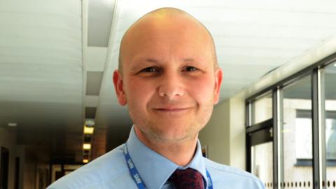 Phil Woodford who is bald with stubble wears a pale blue shirt and red tie and NHS blue and white lanyard. He is standing in a corridor close to windows and smiling.