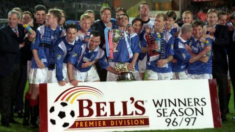 Rangers football team wearing blue shirts and white shorts standing in front of a banner on the pitch that reads BELL'S PREMIER DIVISION WINNERS SEASON 96/97. The team are celebrating and holding two trophies.