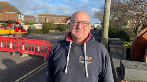 Des Barnett stands next to the A52 roadworks in Mablethorpe. He has a bald head and is wearing glasses. He is wearing a dark hooded top and red polo shirt.