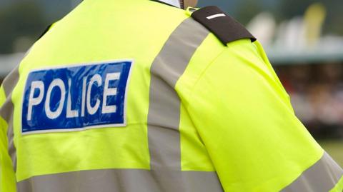 The back of a policeman who is wearing a bright yellow jacket with grey lining. There is a blue panel across the shoulders with the word "police" in white lettering