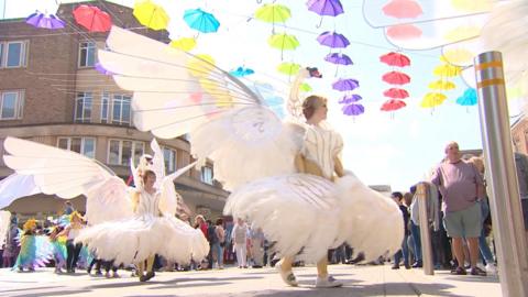 Women dressed in swan costumes parade through the streets. They are wearing huge feathers and have wings. They are surrounded by crowds in the centre of Exeter. Above them, there is an art installation of rainbow coloured umbrellas.