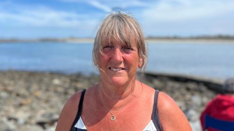 Chrissie Desperques, smiling in her bathers smiling stood at the pier. 

She is a woman in her 50s-60s and has grey/brown hair and blue yes but she is squinting in the sun. Her swimming costume is black and white stripey. 