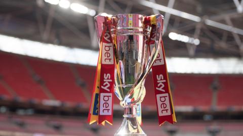 The Championship play-off final trophy. It is silver and there are red, white and yellow ribbons tied around the handles of the trophy that read 'Skybet'. It is standing against the backdrop of Wembley Stadium.
