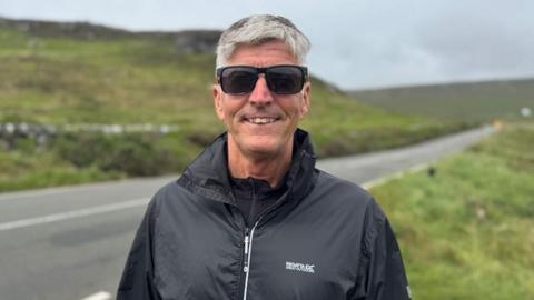 Andrew Titley has short grey hair, is wearing black sunglasses and wearing a black waterproof jacket, stood in front of a road lines with grass.