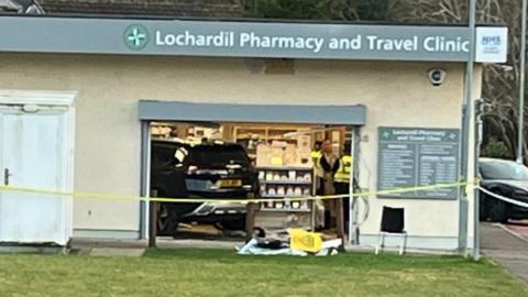 The car is just visible inside the pharmacy. There are police officers in the shop and yellow tape outside the entrance.